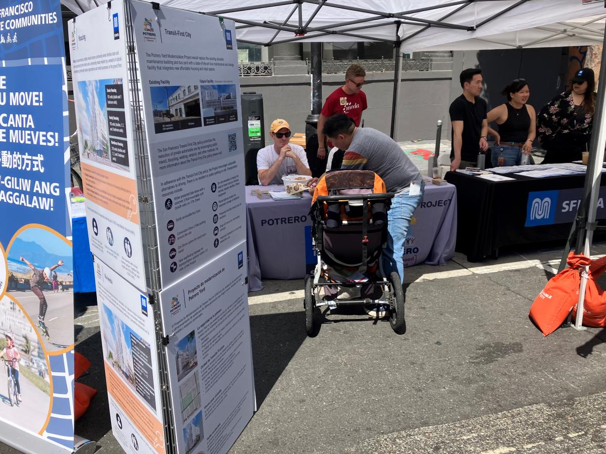 SFMTA staff gathered under white tents at an outdoor event with informational posters about the Project and tables. A man with a baby in a stroller interacts with staff.