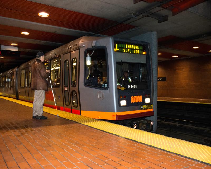 Two passengers waiting for Muni Metro on the platform at Castro Station, one passenger uses a white cane as a mobility too