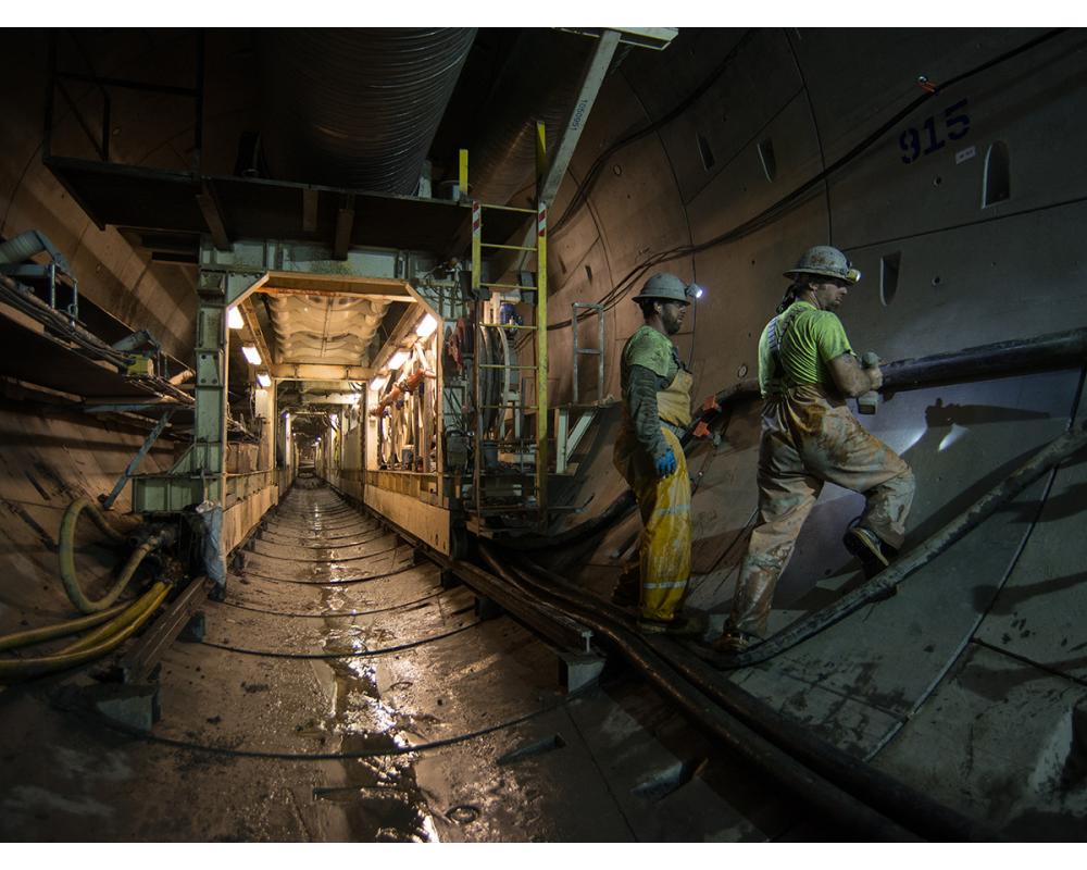 Workers attach new sections of utility pipes and conduit to the southbound tunnel wall as the trailing gear moves forward.