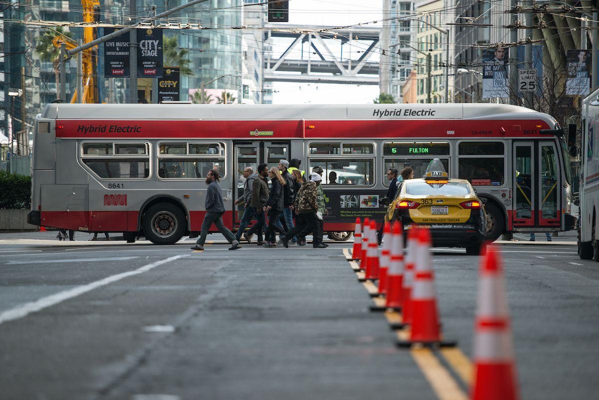 A photograph of the 5 Fulton driving downtown San Francisco, with pedestrians crossing the street alongside the bus.