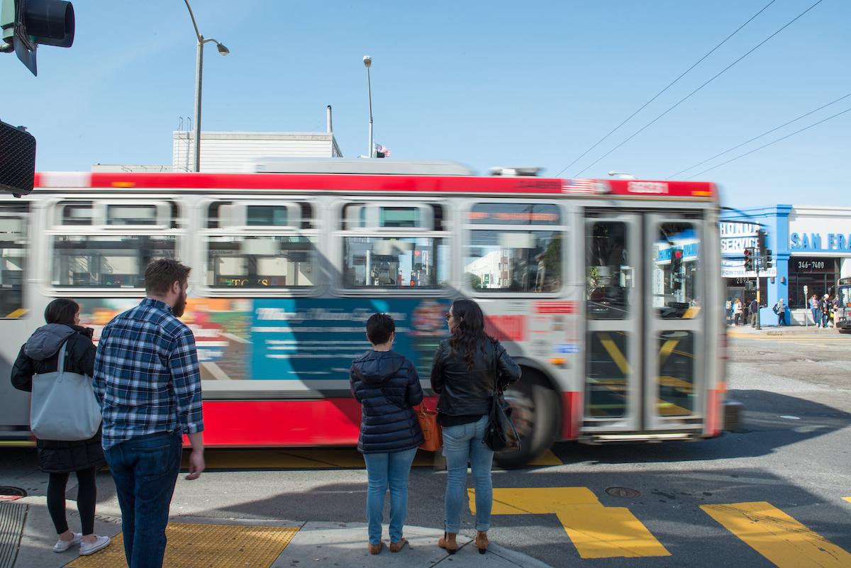 People wait to cross the street as a bus passes in front of them in an intersection.