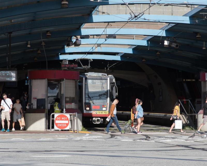 Muni train approaches exit of West Portal Station as people walk in the crosswalk in front of the train.