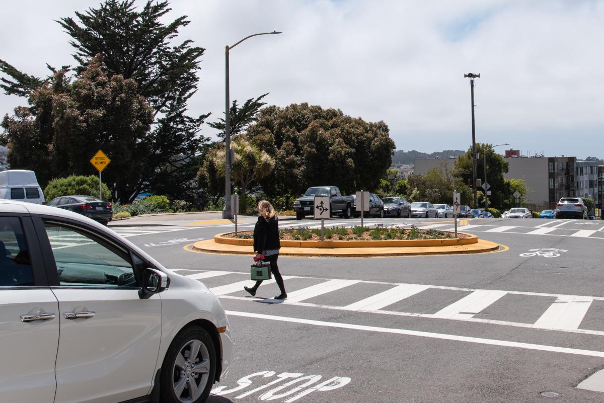 Image shows a traffic circle in the middle of the intersection. A vehicle is yielding to a pedestrian who is crossing in the crosswalk.