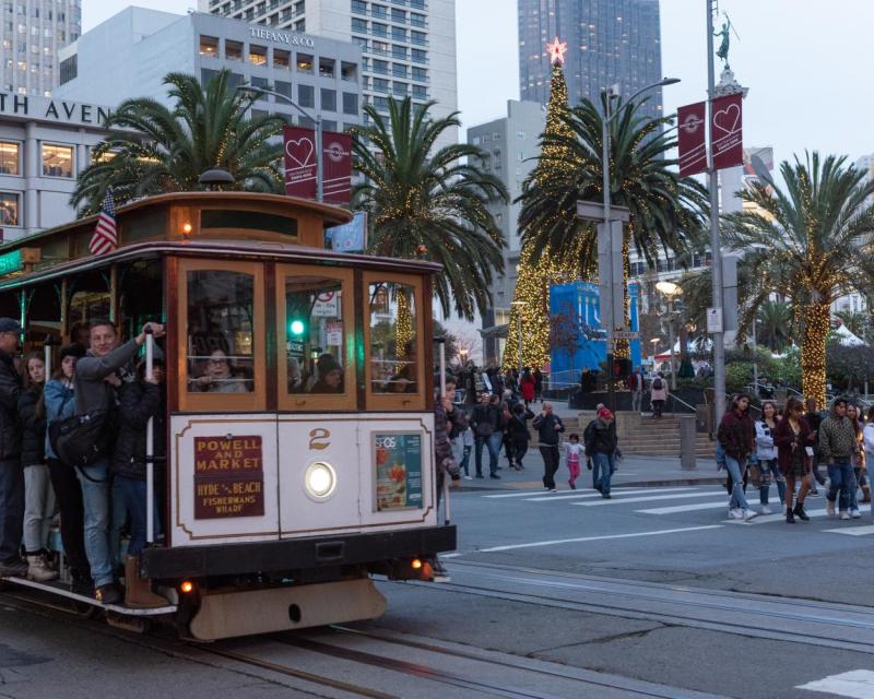 Photo of Union Square with holiday lights in the backgrouns, people crossing the street, and people riding a Cable car in the fo