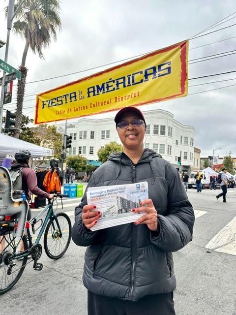  A person in a black jacket and cap holds a brochure while standing at an outdoor event. A banner with the text "Fiesta de las Américas" spans the street in the background.