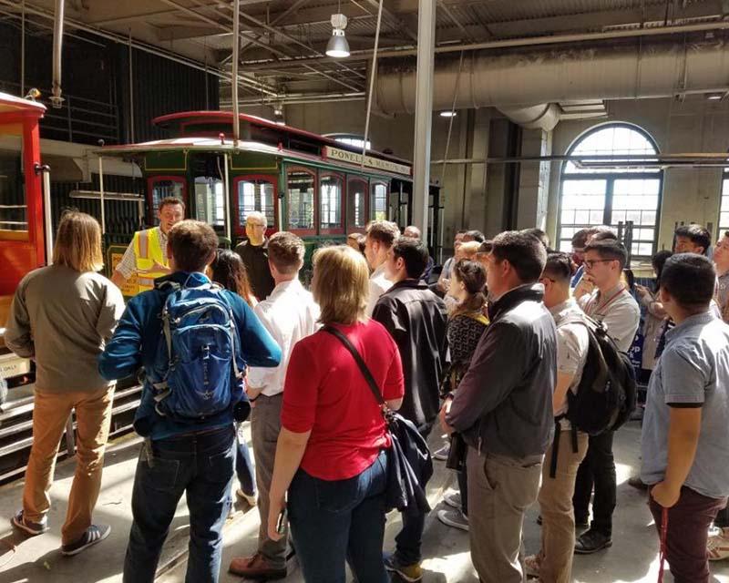 Interns at cable car barn; SFMTA employees and a cable car in the background