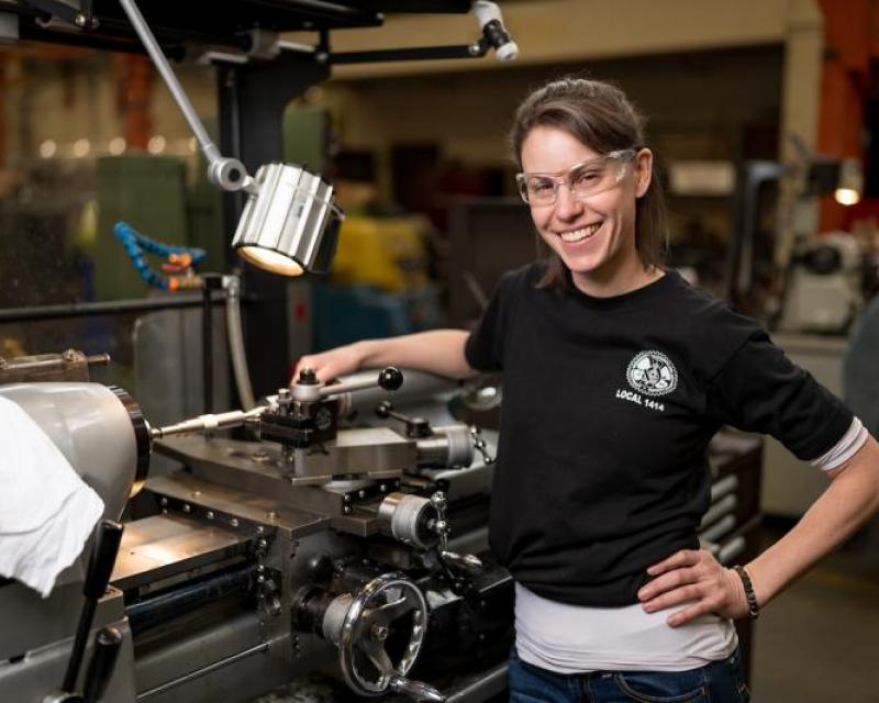 Brittany McMartin smiles over a lathe. We see a range of lights and other machinery around her setup.