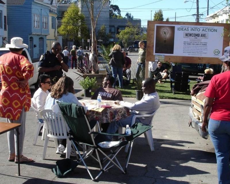 People gather at tables at a street meeting. Poster reads "Ideas into action".