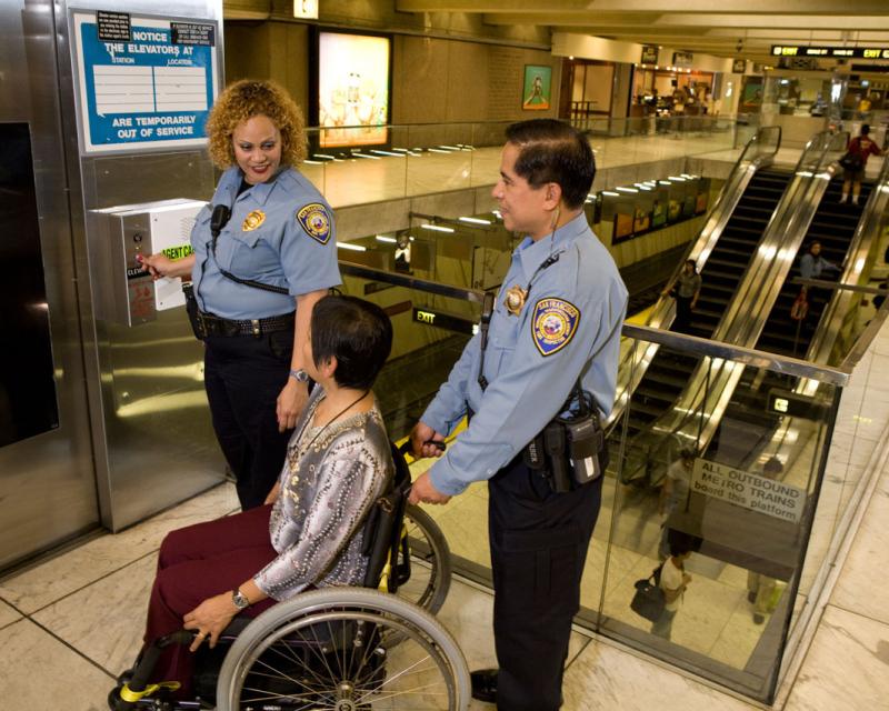 Image of two Muni safety officers helping a woman in a wheelchair into an elevator