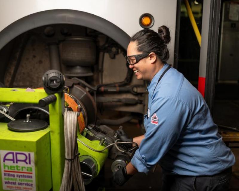 Jenny measures break rotors for a vehicle. We see her smiling and wearing a blue uniform as she works on a green machine.