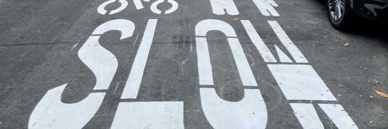 Slow Streets pavement markings in white show the outline of a cyclist, an adult and a child, and the word SLOW.