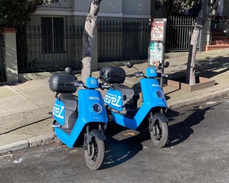 Two shared electric mopeds parked at the curb on a residential street.