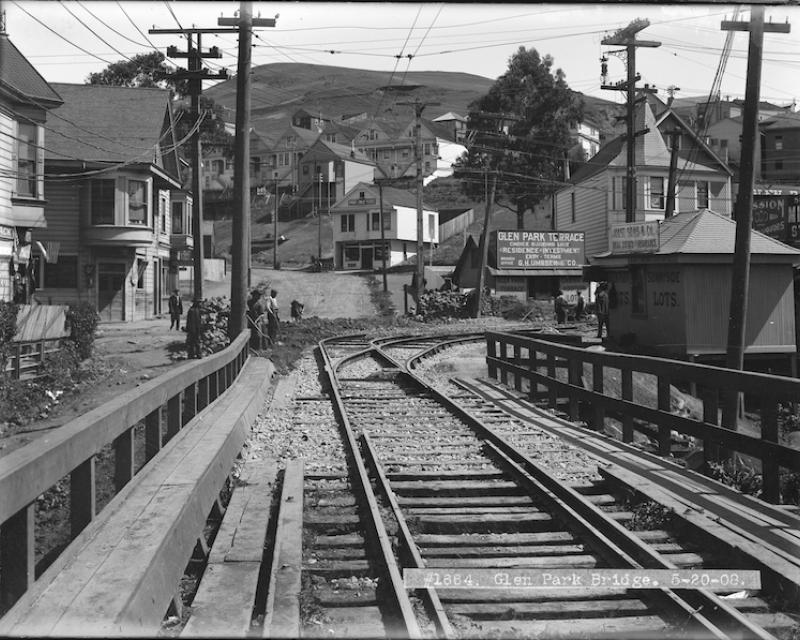 Black and white photo showing a view north on Diamond Street near Chenery in San Francisco's Glen Park Neighborhood. in the foreground is a wooden bridge and streetcar rails, followed by a street curving uphill with a handful of early 20th century wooden houses and buildings.