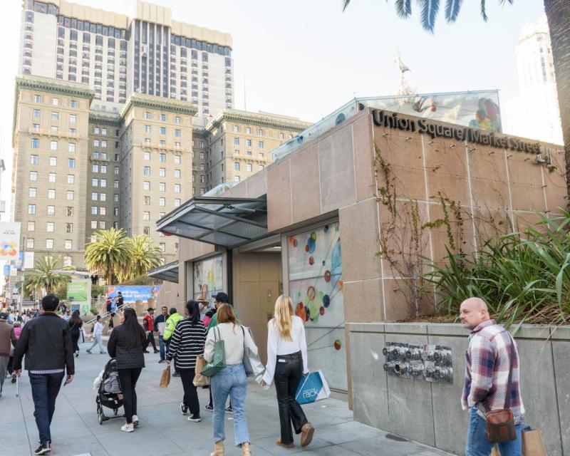 Street view of a station entrance and downtown buildings with dozens of pedestrians holding various items 