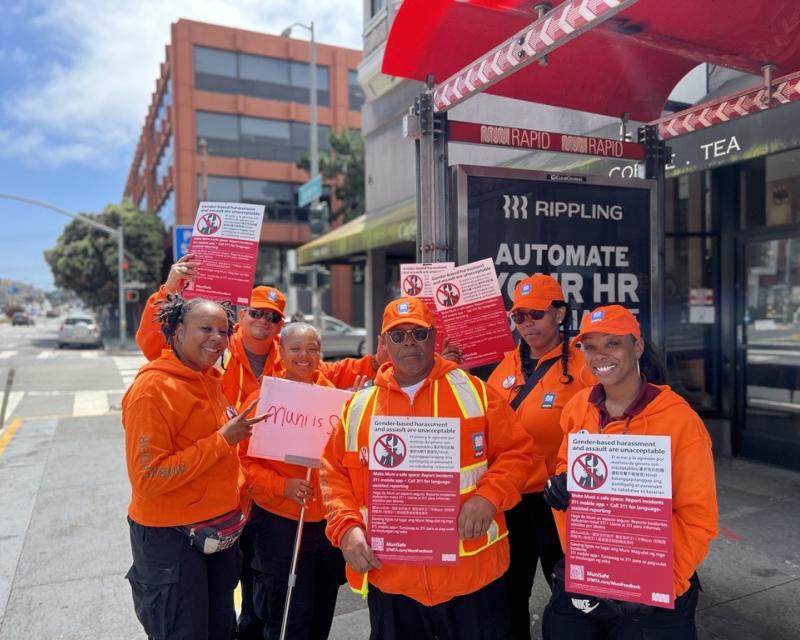 Group of people at a Muni bus stop holding signs about reporting harassment on Muni